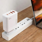A white Dudao 65W GaN fast charger is plugged into a power strip on a wooden table, connected by a gray cable to a silver laptop nearby. The charger has multiple ports (USB-C1, USB-C2, USB-A1, USB-A2) and "GaN" and "65W" labels visible on its side.